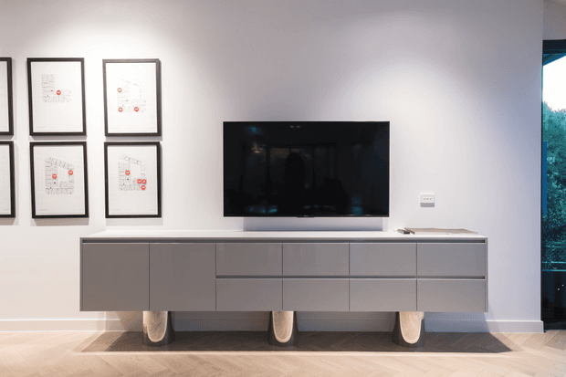 Contemporary media wall with a wall-mounted flat-screen television above a long, minimalist grey built-in or freestanding storage console. The white wall is decorated with a grid of black-framed prints, while the light timber herringbone floor and dark rug add warmth and contrast. A tall glazed opening at right provides a glimpse of greenery outside.