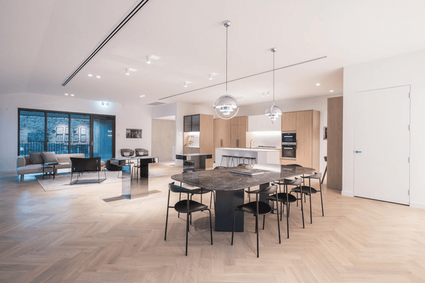 A spacious open-plan contemporary interior combining a kitchen, dining area and living room. The room features pale herringbone timber flooring, light timber kitchen cabinetry, a dark stone island and dining table, black-framed glazing, minimalist black dining chairs, and globe pendant lights. A lounge area is visible at the rear beside large glazed doors.