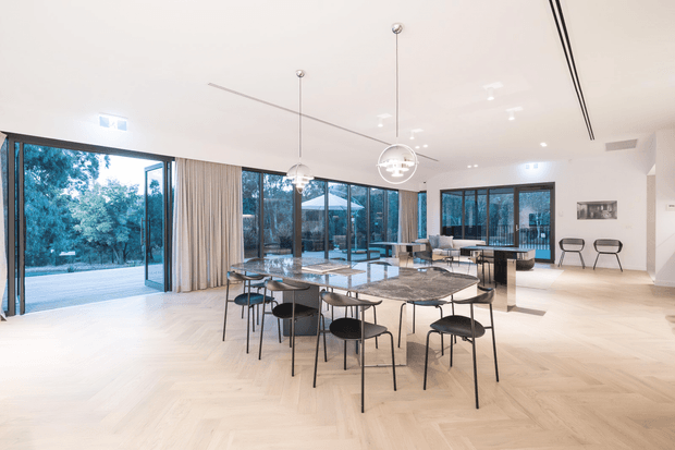 A bright, spacious contemporary open-plan dining and living area with floor-to-ceiling black-framed glazing on multiple sides, opening toward a landscaped outdoor setting. A large stone dining table with black chairs occupies the foreground, while a lounge seating area and a dark kitchen island or bar are visible beyond. Pale herringbone timber flooring, sheer curtains, sculptural pendant lights, and restrained neutral furnishings create an airy, refined interior.