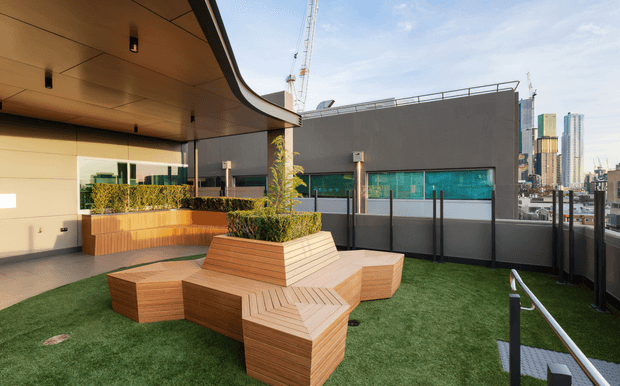 A contemporary rooftop terrace with artificial grass, built-in geometric timber seating arranged around a raised planted timber planter, and a curved covered canopy with recessed lighting. A dark parapet wall, translucent privacy screening, construction cranes, and a dense high-rise city skyline are visible beyond.