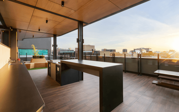 A spacious rooftop terrace with a contemporary outdoor kitchen and bar area, featuring dark built-in cabinetry, a large island or counter, warm timber ceiling panels, timber-look flooring, black-framed structural posts, and glass balustrades. The terrace overlooks a dense urban skyline in warm late-afternoon light. A built-in planter and outdoor seating are visible in the background.