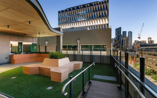 A contemporary rooftop terrace with artificial grass, angular timber-clad built-in seating, stainless steel handrails, and a gently curved roof canopy. The terrace overlooks a dense urban skyline with high-rise buildings and construction cranes. A distinctive patterned screen or facade element rises above the adjacent building.