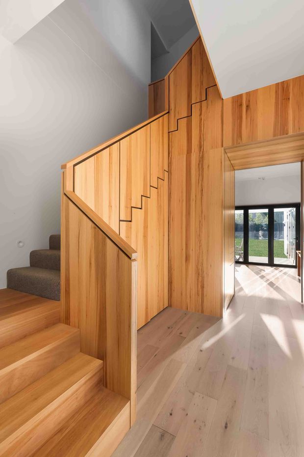 Bright contemporary interior view of a timber-lined stairway and hallway. The staircase has warm vertical timber wall panelling, matching timber treads, a simple timber handrail, and carpeted upper steps. Pale timber flooring continues through the hall toward a glazed living area, with strong daylight entering from black-framed doors and windows.
