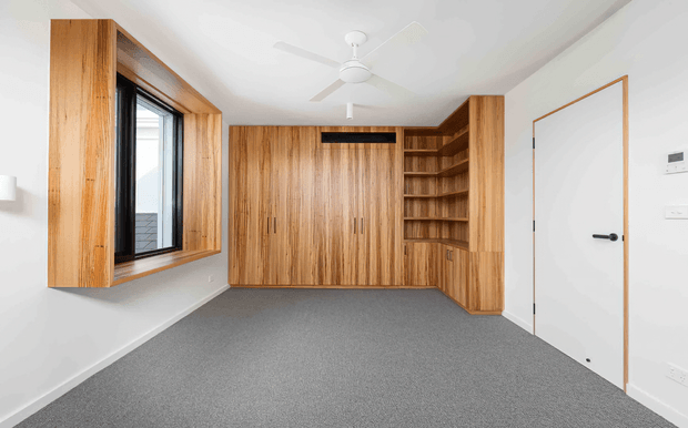 Bright, mostly empty bedroom or study with extensive custom timber built-in cabinetry, open shelving, a desk-height cabinet section, and a window seat surround. The room has white walls and ceiling, grey carpet, a white ceiling fan, black-framed glazing, and a white internal door with timber trim.