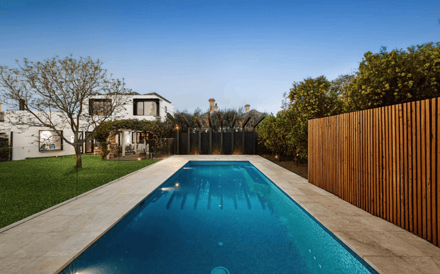 A long rectangular swimming pool occupies the foreground of a landscaped residential backyard, bordered by pale stone paving. A modern white house and a darker gabled structure sit beyond the pool, with mature trees, lawn, shrubs, and a tall vertical timber privacy fence enclosing the space. The image is a clear, sunny exterior architectural photograph.