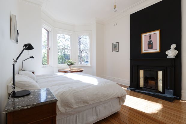 Bright bedroom in a period-style home, featuring a large white upholstered bed, polished dark stone-topped bedside furniture, tall bay windows, timber floorboards, and a striking black-painted fireplace wall with a traditional mantel and framed artwork. Natural light creates a warm, refined atmosphere.