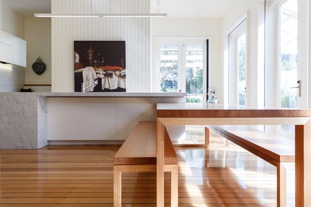 Bright open-plan kitchen and dining area with white cabinetry, a long stone island bench, vertical white wall panelling, and extensive glazing. A built-in timber dining table and bench seating extend from the island, coordinating with the warm timber floor and furniture. The space has a clean, contemporary architectural character with abundant natural light.