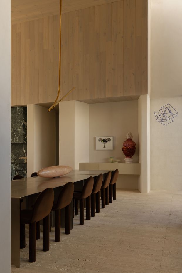 A warm, refined dining area with a long stone dining table and a row of dark upholstered dining chairs. The room features extensive light timber wall and ceiling panelling, a minimalist built-in sideboard, pale stone flooring, sculptural pendant lighting, and restrained decorative objects. A dark stone-clad opening and geometric wall artwork add contrast.
