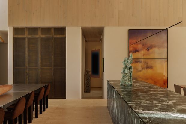 Warm, refined open-plan dining and living interior with extensive light timber wall and ceiling panelling. A dark timber built-in cabinet occupies the left wall beside a dining table with upholstered chairs. In the foreground, a substantial stone-topped island or console features a dramatic veined dark surface and textured stone-clad base. Neutral plaster walls, sculptural decor, and a large abstract artwork complete the contemporary space.