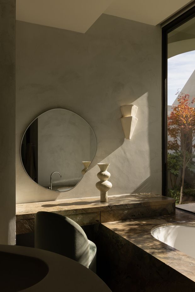 A softly lit, sculptural bathroom vanity area with a large round mirror, integrated stone vanity and basin, curved dark upholstered chair, decorative vessel, and sculptural wall light. Warm natural light enters through full-height black-framed glazing, creating dramatic shadows across the textured plaster walls and stone surfaces.