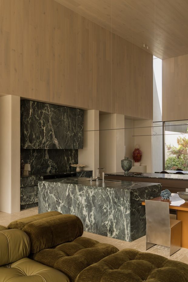 A sophisticated open-plan kitchen and dining area viewed from a living room. The space features extensive warm timber wall and ceiling lining, a dramatic dark green veined stone island and matching splashback, integrated cabinetry, sculptural dining furniture, and a large olive-brown upholstered sectional in the foreground. Full-height glazing brings in daylight and connects to planted greenery outside. The design is luxurious, warm, and highly material-focused.