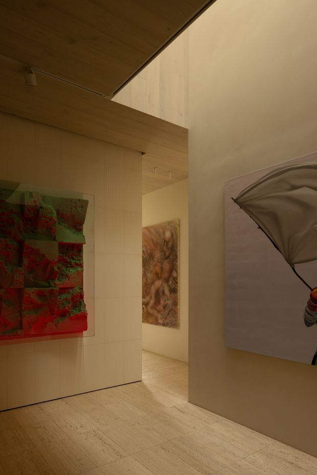 A warm, softly lit contemporary interior gallery or hallway with pale wood flooring, wood-clad ceiling planes, and light neutral partition walls. Several large abstract artworks are displayed on the walls, while concealed or minimal lighting creates a refined, intimate atmosphere. The view opens toward another artwork-lined passage in the background.