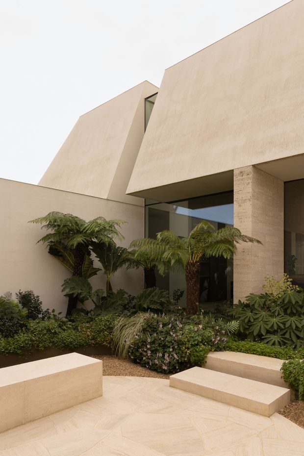 A contemporary residential courtyard viewed from an outdoor terrace, featuring sculptural angular beige rendered walls and roof forms, large dark-framed glazing, and a textured stone or concrete column. The foreground has pale geometric paving and built-in beige seating blocks, while lush fern and shrub planting softens the minimalist architecture.