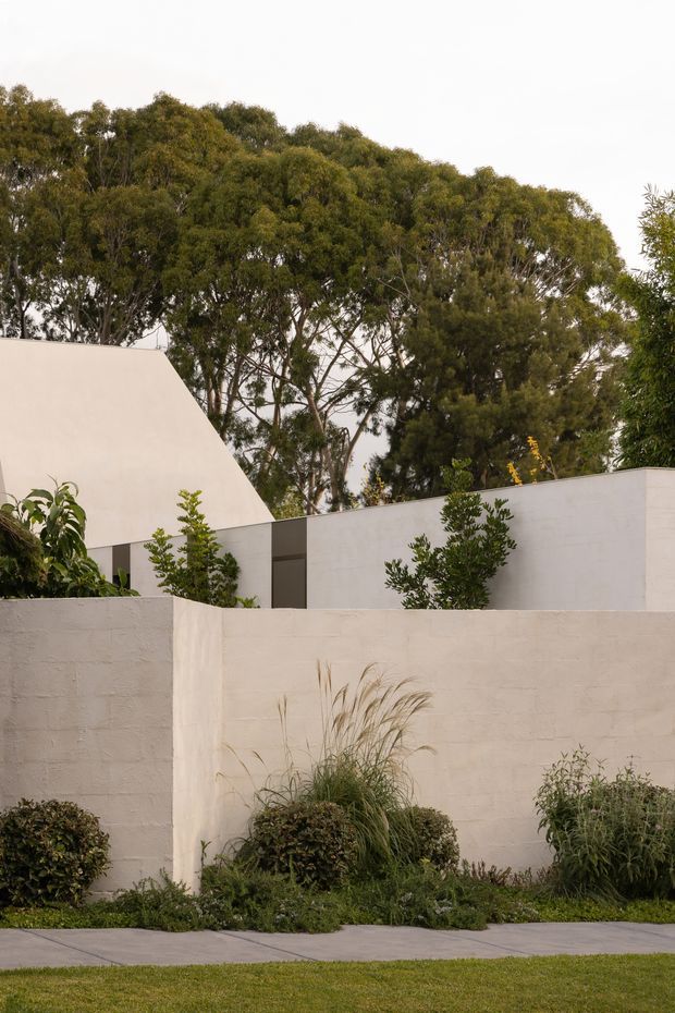 Exterior view of a low, minimalist residential building partly screened by a pale rendered masonry boundary wall. The composition features clean geometric volumes, a steep angular roof form at left, muted grey-white surfaces, simple dark-framed openings, and layered ornamental planting with mature trees behind.