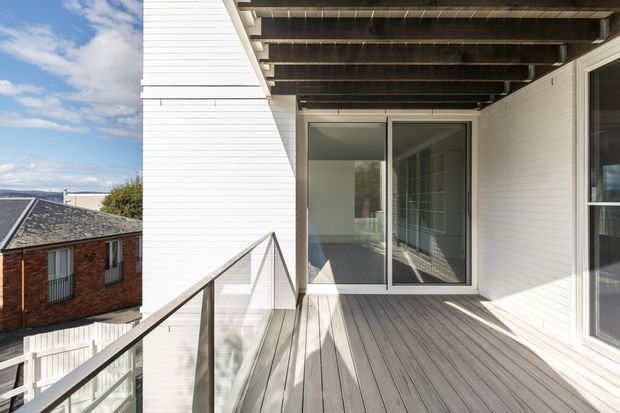 A bright contemporary exterior balcony or terrace viewed from outside a white-clad building. The space has grey timber decking, a slim dark metal and clear-glass balustrade, white horizontal wall cladding, exposed timber beams overhead, and large sliding glass doors leading into a simple interior. Nearby residential rooftops and a glimpse of the coastal horizon are visible beyond the balcony.
