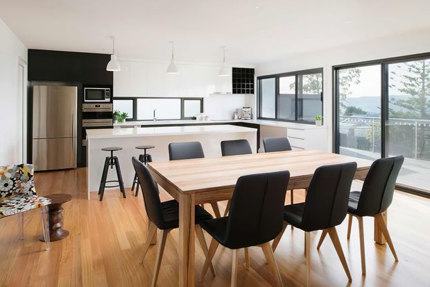 Bright open-plan kitchen and dining area with a contemporary timber dining table and black upholstered chairs in the foreground. The kitchen features white cabinetry, a dark contrasting wall and island base, stainless-steel appliances, pendant lighting, and timber flooring. Large black-framed windows and sliding glazing provide views toward a body of water and surrounding landscape.