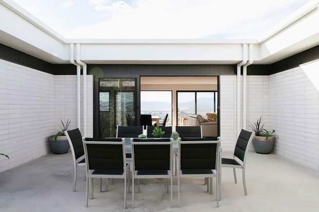 A bright, modern courtyard terrace with a flat-roofed open-air enclosure. White tiled walls frame a dining setting with a rectangular table and six black-and-white outdoor chairs. Large glazed doors open toward an interior living area, while potted plants soften the minimalist hardscape. The space has a light, airy contemporary feel and appears designed as an outdoor dining and entertaining area.