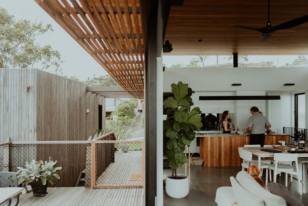 A contemporary indoor-outdoor entertaining area viewed from a timber deck. The covered space features a slatted timber pergola and ceiling, large openings, a timber kitchen island with stools, white dining furniture, a lounge area, planted greenery, and a fenced courtyard beyond. The scene has a relaxed, warm modern character with strong visual continuity between the interior and exterior.