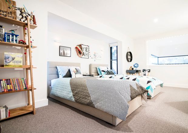 Bright, spacious bedroom with two beds, upholstered grey headboards, layered blue, white and grey bedding, and a large beige carpet. The room features white walls and ceiling, a narrow vertical window, a long horizontal window, simple framed artwork, and a tall open timber shelving unit filled with books and decorative items. The overall look is clean, airy, and contemporary with playful geometric accents.