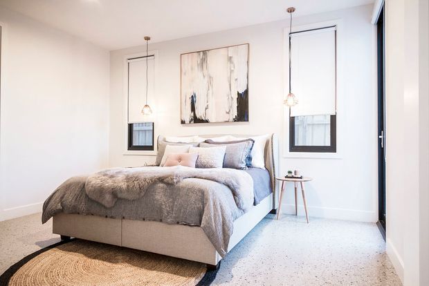 Bright, softly styled bedroom with a large upholstered bed dressed in neutral grey and blue bedding, two narrow black-framed windows, pendant lights, a small timber side table, abstract wall art, pale walls, and light carpet. The room has a clean, calm, contemporary feel with warm natural accents.