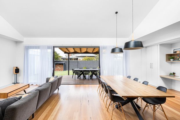 Bright open-plan contemporary living and dining area with pale timber flooring and a large timber dining table seating many people. A grey sectional sofa and timber media console occupy the living zone, while white sheer curtains frame wide glazing and an outdoor dining area beyond. White built-in cabinetry and open shelving line the right wall, with three black pendant lights above the dining table. The room opens to a covered outdoor terrace or courtyard.