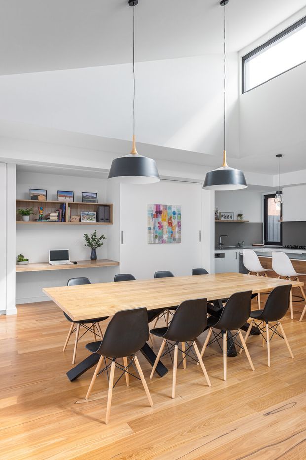 Bright open-plan dining and kitchen interior with a long light timber dining table surrounded by black and white molded chairs. The space has warm timber flooring, white walls and ceiling, a built-in timber study nook and shelving, contemporary pendant lights, colorful wall art, and a compact kitchen with grey cabinetry and a peninsula visible in the background. High-level glazing brings in abundant natural light.