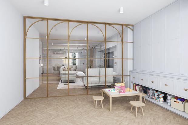 Bright, refined children's playroom with pale herringbone timber flooring, white decorative wall panelling and extensive built-in cabinetry. A gold-toned framed glass partition with arched detailing separates the play area from a sophisticated adjoining living or dining space. The room contains a low children's table and stools, toys, and built-in open storage, with a calm neutral palette and brass-gold accents.