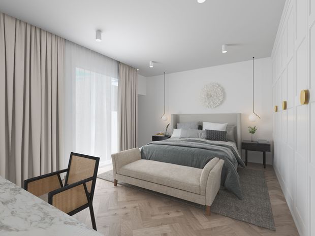 A refined contemporary bedroom with a large upholstered bed, layered grey bedding, a curved beige bench at the foot of the bed, and soft full-height curtains covering broad glazing. The room features light herringbone timber flooring, a grey area rug, a white paneled built-in wardrobe wall with small brass hardware, minimalist pendant bedside lights, and a marble-look surface with dining-style chairs in the foreground. The overall palette is light, neutral, and softly textured.