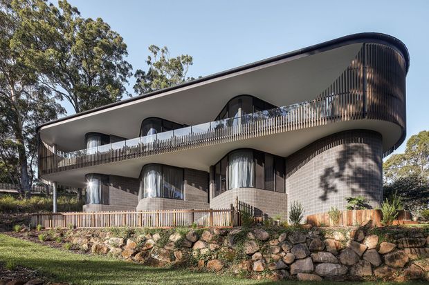Exterior view of a contemporary multi-storey residential building on a landscaped, sloping site. The design features a strongly curved upper roof and balcony edge, rounded glazed corners, continuous balconies with slim vertical balustrades, dark window frames, and pale textured masonry walls. Natural stone retaining walls and mature trees surround the building.