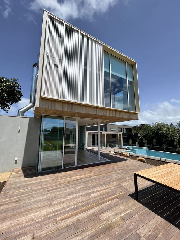 Exterior view of a contemporary multi-level residence with a projecting upper storey, extensive floor-to-ceiling glazing, vertical white screening fins, and timber cladding beneath the cantilever. A broad timber deck surrounds the house and leads to a rectangular swimming pool. The scene is photographed from the outdoor deck under a bright blue sky.