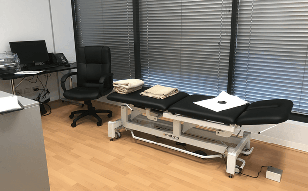 A compact professional treatment or examination room with a black adjustable treatment table centered in front of large windows fitted with horizontal venetian blinds. A black office chair and workstation with computer equipment sit to the left, while folded towels and paperwork rest on the treatment table. The room has pale timber flooring, white walls, and a clean, functional clinical fit-out.