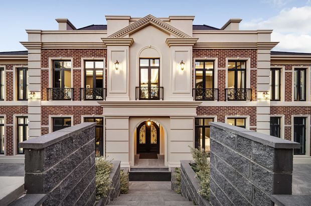 Front exterior view of a grand multi-storey residential building with symmetrical classical composition, red brick wings, pale rendered central entrance pavilion, ornate triangular pediment, pilasters, decorative cornices, tall multi-pane windows, wrought-iron Juliet balconies, and a formal arched front door. Dark stone boundary walls and steps lead toward the entrance.
