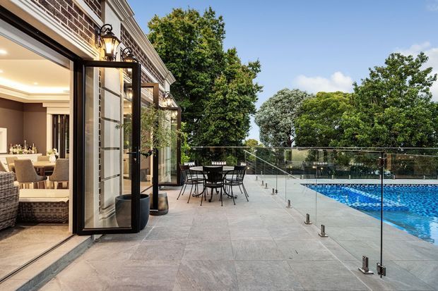 A refined outdoor terrace beside a swimming pool, adjoining a brick-clad home through large black-framed bi-fold or hinged glazed doors. The terrace is finished in large pale stone tiles and enclosed by a frameless glass pool fence, with outdoor dining furniture, potted plants and traditional wall-mounted lanterns. The open doors reveal a warmly styled dining and living interior.