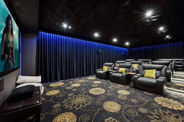 A dark, luxurious home cinema with multiple rows of large black leather reclining theater seats, bright yellow accent cushions, blue LED lighting washing over pleated dark curtains, a patterned carpet, recessed ceiling spotlights, and a large projection screen on the left wall. The room has an immersive, moody entertainment-focused atmosphere.