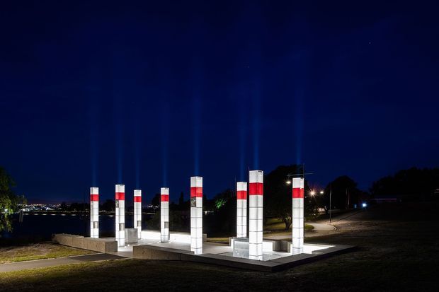 Night-time exterior view of a landscaped public or institutional site featuring a geometric illuminated installation of tall rectangular tiled columns, accented with red and dark horizontal bands. The installation sits on a raised paved platform beside a waterfront or dark open landscape, with pathways, trees, and distant lights visible in the background.