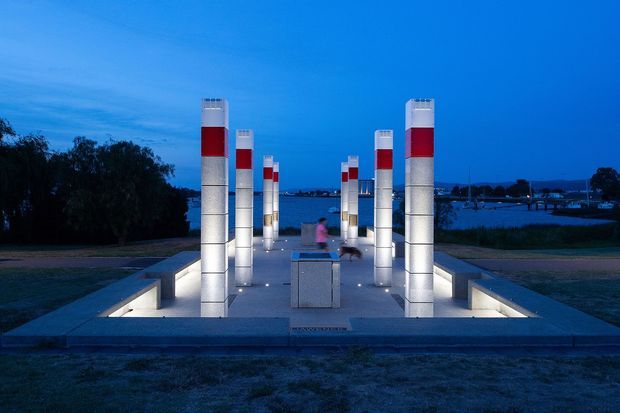 A dusk exterior view of a formal illuminated outdoor installation or public space beside a waterfront. Symmetrical rows of tall rectangular light columns, patterned with white and red sections, frame a central paved axis and square plinth. Low linear walls with integrated lighting define the foreground, with water and distant shoreline visible beyond.