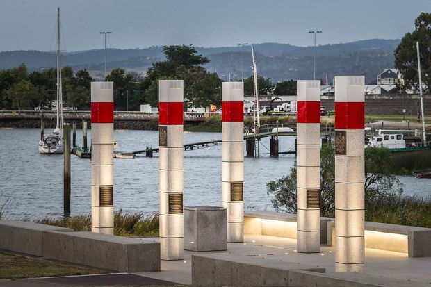 A waterfront public promenade or landscaped architectural installation featuring five illuminated rectangular stone-clad columns with bold red bands and inset bronze plaques. Concrete seating and retaining elements frame the foreground, while a marina, boats, harbour infrastructure, and wooded hills are visible beyond. The scene is photographed at dusk with integrated ground and column lighting.