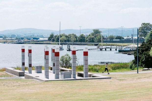 An outdoor waterfront public space with a geometric sculptural installation made of tall rectangular grey columns accented with red and white sections. The installation sits on a paved platform beside a broad body of water, with a pedestrian path, grassy open lawn, marina infrastructure, boats, trees, and distant hills in the background.