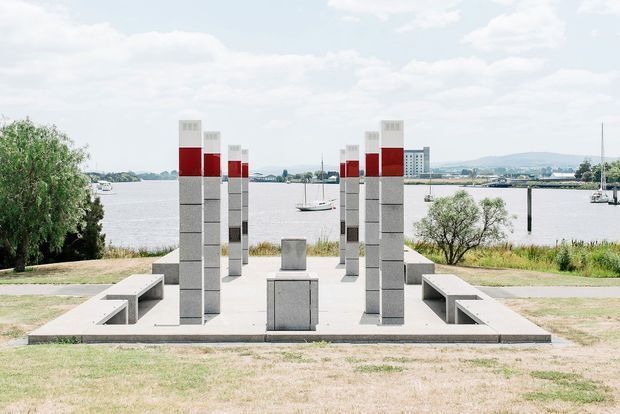 A symmetrical waterfront memorial or civic installation set on a paved platform, with rows of tall rectangular grey stone or concrete pillars featuring red and white upper sections. Low built-in benches flank a central pedestal, while a river, boats, distant buildings, trees, and hills form the background under a bright partly cloudy sky.