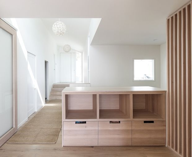 Bright, minimalist residential hallway or entry area with pale timber built-in cabinetry forming a low room divider, open shelving above drawers, and a matching vertical timber-slat feature wall. The space has white walls, light timber flooring, beige carpeted stairs, white paneled doors, a small square window, and delicate spherical pendant lights. The composition looks toward a stair landing and entry circulation zone.