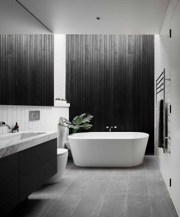 A contemporary bathroom featuring a freestanding white bathtub, wall-mounted black vanity with a marble benchtop, grey floor tiles, white rectangular wall tiles, and a dramatic black vertical timber feature wall. A small plant and heated towel rail add functional and natural accents.