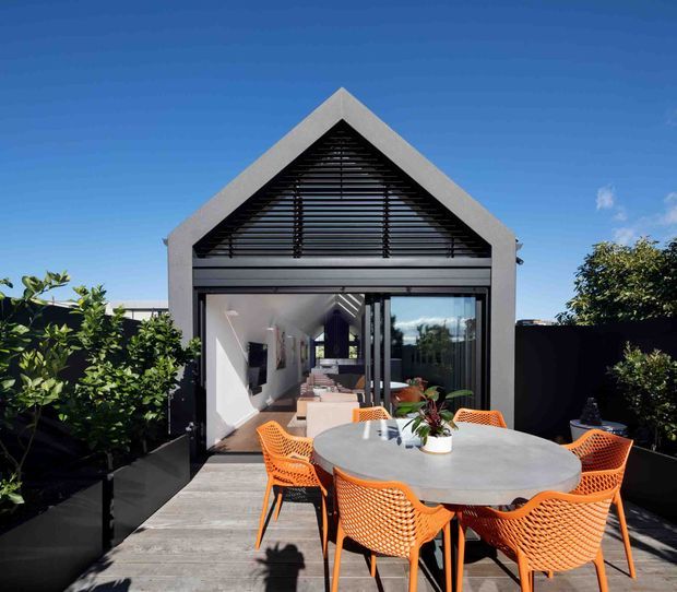 Exterior view of a contemporary gabled pavilion or rear extension opening onto a timber deck. The facade features a dark-framed glazed sliding opening, a prominent triangular gable with horizontal louvres, and a round outdoor dining table with bright orange chairs in the foreground. Hedging and planters frame the deck under a clear blue sky.