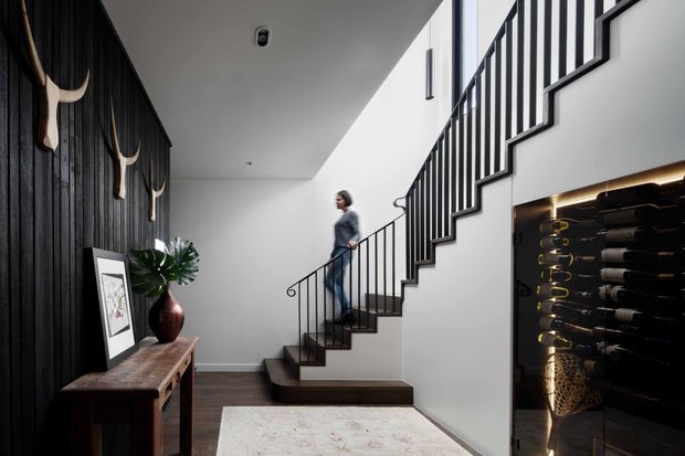 A refined modern residential entry hall featuring a dark vertical timber feature wall, console table, framed artwork, sculptural wall decor and indoor plant. A black metal stair balustrade rises beside white walls, while an illuminated built-in wine display is visible beneath the staircase. Dark timber flooring and a light area rug create contrast.