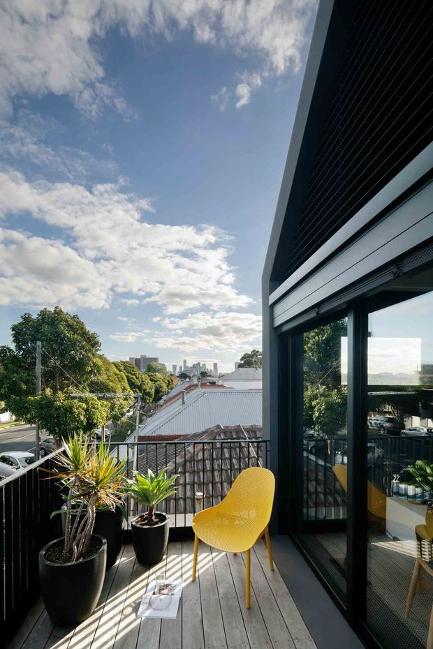 A sunny elevated balcony or terrace with grey timber decking, black metal balustrades, potted architectural plants, and a bright yellow outdoor chair. Large dark-framed sliding glass doors open to the contemporary building, reflecting the terrace and surrounding urban neighbourhood. Rooftops, trees, and a distant city skyline are visible beyond.