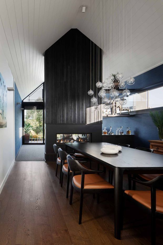 A dramatic dining room with a long dark dining table and black-framed chairs with tan leather seats. The room features dark blue walls, a tall black vertical timber-clad architectural volume with an integrated fireplace, white painted timber-lined pitched ceilings, dark timber flooring, and a cluster of suspended glass globe pendant lights. Glazing and a glazed door open toward a leafy outdoor area.