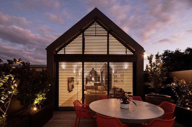 Dusk exterior view of a compact contemporary pavilion-like house or entertaining space, with a steep gabled roof, full-height glazed facade screened by horizontal louvres, and a timber deck furnished with a round dining table and red outdoor chairs. Planting and subtle landscape lighting frame the structure, while a warm, softly lit interior is visible through the glazing.