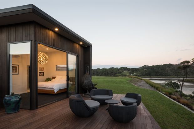 A compact modern pavilion or bedroom retreat with dark vertical timber cladding, a flat roof, and extensive glass doors opening onto a timber deck. The interior visibly contains a bed and bedside furnishings, while the deck has a small arrangement of dark outdoor lounge chairs and tables. The building sits beside a landscaped lawn and calm waterside setting, photographed at dusk.