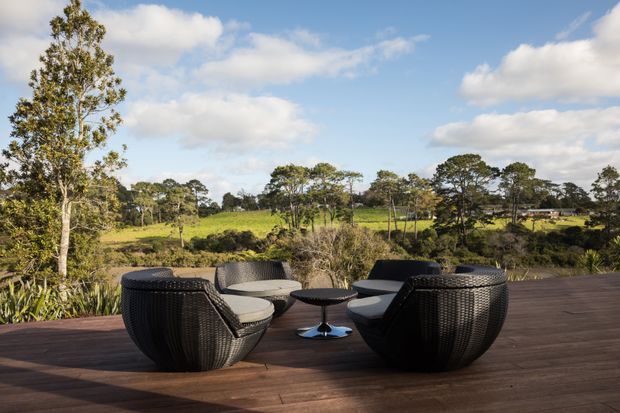 An outdoor seating area on a broad timber deck, furnished with dark woven lounge chairs, light cushions, and a small round pedestal table. The deck overlooks a green rural landscape with scattered trees under a bright partly cloudy sky.