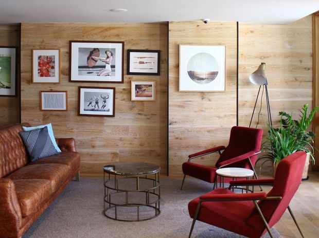 A warm, mid-century-inspired living room with full-height timber wall panelling, a brown leather sofa, red upholstered lounge chairs, a black metal tiered coffee table, framed artwork, and indoor greenery. The room has a low, relaxed seating arrangement and a neutral carpeted floor.