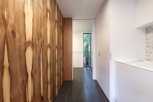 A narrow, contemporary hallway with dramatic full-height natural timber built-in cabinetry along the left wall and smooth white built-in cabinetry on the right. The dark large-format floor leads toward a glazed doorway with greenery visible beyond. A partial white benchtop and small tiled splashback are visible at right, suggesting an adjacent kitchen or utility area. The space is bright, clean-lined, and materially contrasting, with standard ceiling height.